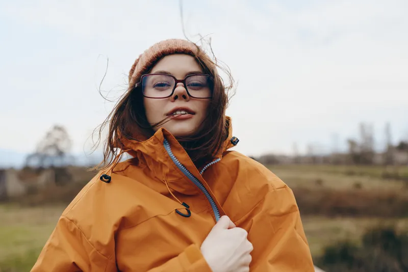 Woman outdoors, orange jacket, glasses, field, wind, portrait, lifestyle concept, standing in a countryside landscape, soft daylight, natural colors, calm mood