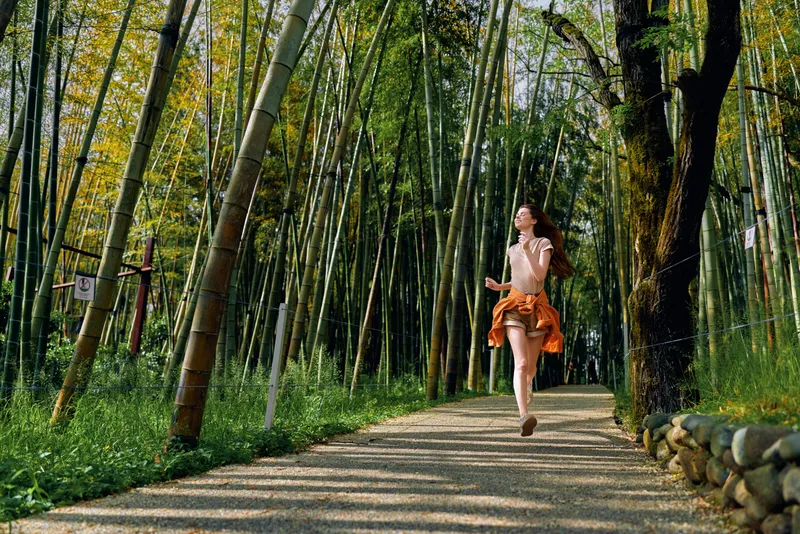 Woman running on a forest trail and gravel path through dense bamboo, wearing a flowing dress as sunlight and motion meet nature in a peaceful outdoor fitness and leisure scene.