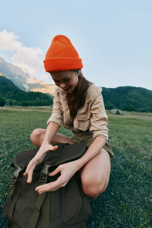 Cheerful woman outdoors, sitting in a grassy meadow by mountains, unpacking a backpack, wearing an orange beanie, relaxed pose, natural light, lifestyle moment