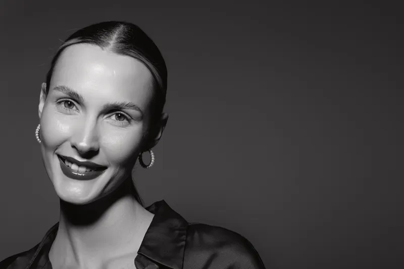 Smiling caucasian woman with light skin and slicked back hair wears hoop earrings and dark shirt. Black and white studio portrait with soft lighting and confident expression.