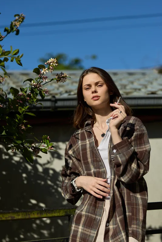 A woman outdoors in a plaid coat stands in a sunlit garden, smoking a cigarette, relaxed and confident. A casual lifestyle portrait in a contemporary urban setting