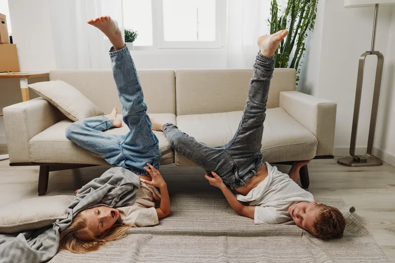 Two children lying on the floor with legs raised, playing video games at home in casual clothes. Bright living room with sofa and natural light creates a cozy atmosphere.
