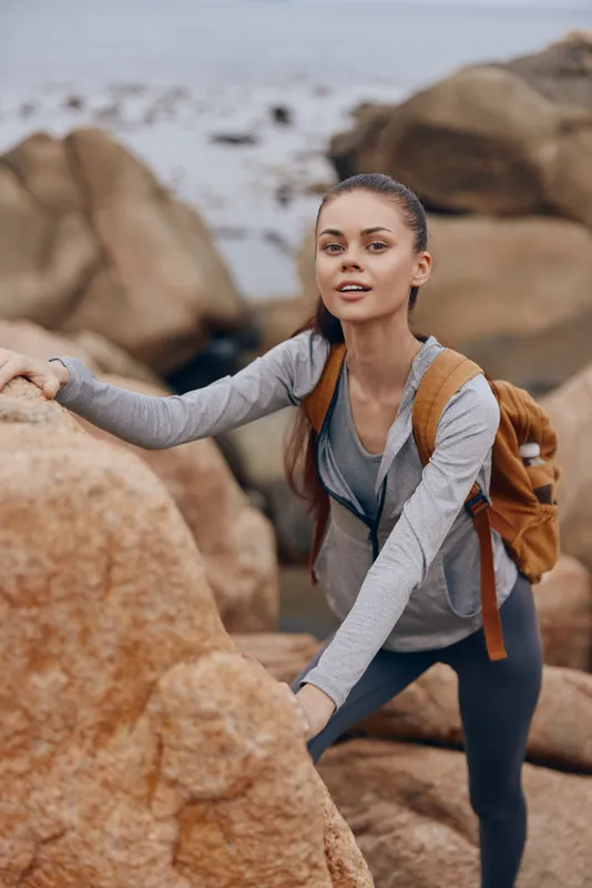 Young female hiker climbing rocks, wearing a gray sports jacket and backpack, with a joyful expression in a natural outdoor setting, showcasing adventure and fitness
