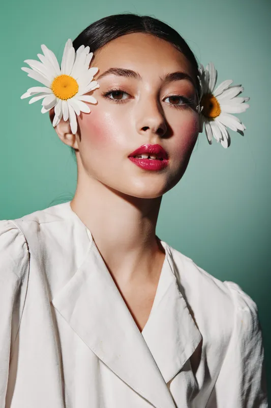 Close-up portrait of a young woman with glossy lips and radiant skin wearing white flowers in her hair against green background fashion beauty editorial concept.