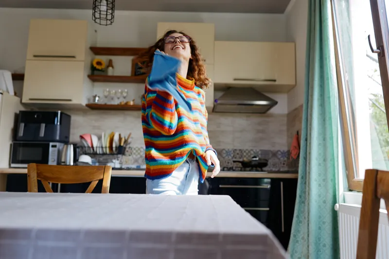 Young woman in a colorful striped sweater stands in a bright kitchen, lifting a blue shirt toward her face with a playful, candid mood.