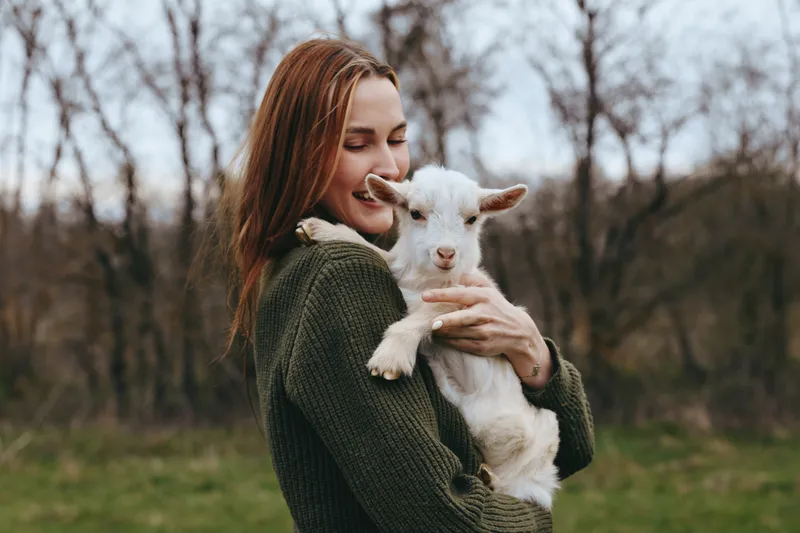 A young woman in a green sweater gently holds a white baby goat outdoors in a natural setting during early spring, showing affection and calm connection with the animal.