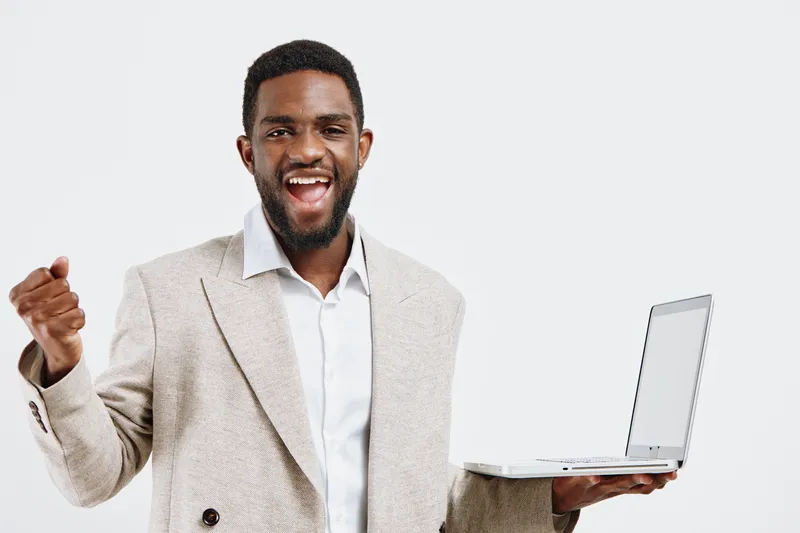 Happy young African American man in beige blazer and white shirt celebrating success while holding open laptop isolated on plain white background. People achievement concept