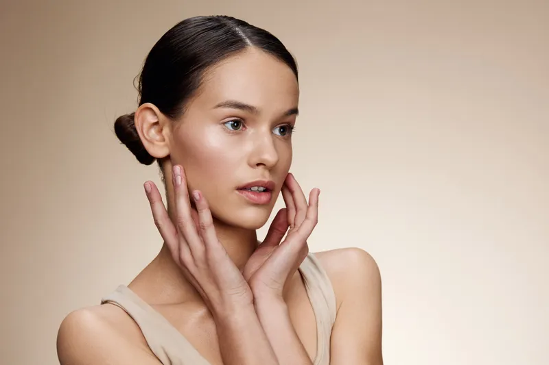 A young woman with a natural look, showcasing healthy skin and minimal makeup, set against a soft beige background, emphasizing beauty and skincare concepts
