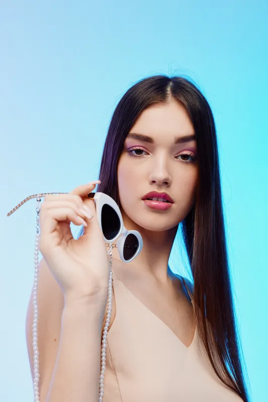 Young woman with long hair holds white sunglasses close to camera. She wears natural makeup and a beige top against a soft blue background. Concept of beauty fashion style.