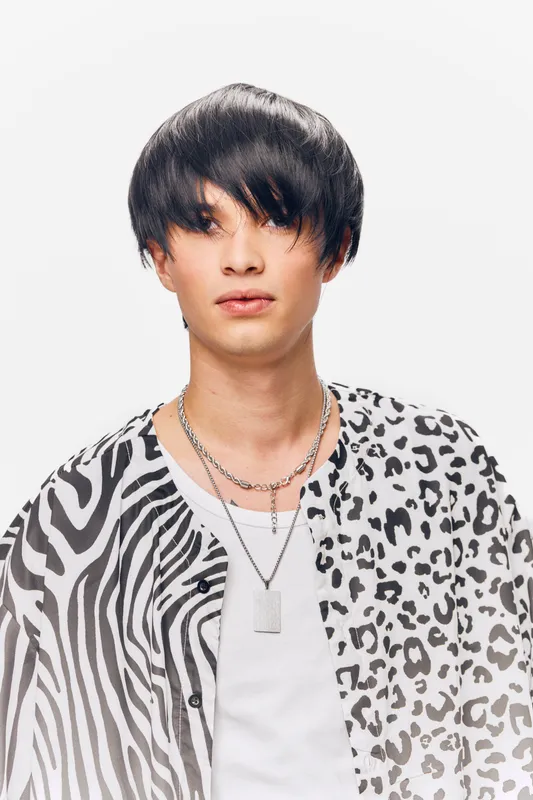 young man wearing a black and white animal print shirt with layered necklaces, minimal makeup, and a simple white background in a studio setting
