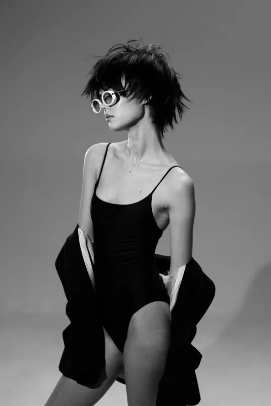 Black and white portrait of a young woman wearing sunglasses and a black swimsuit with a jacket draped over her arms.