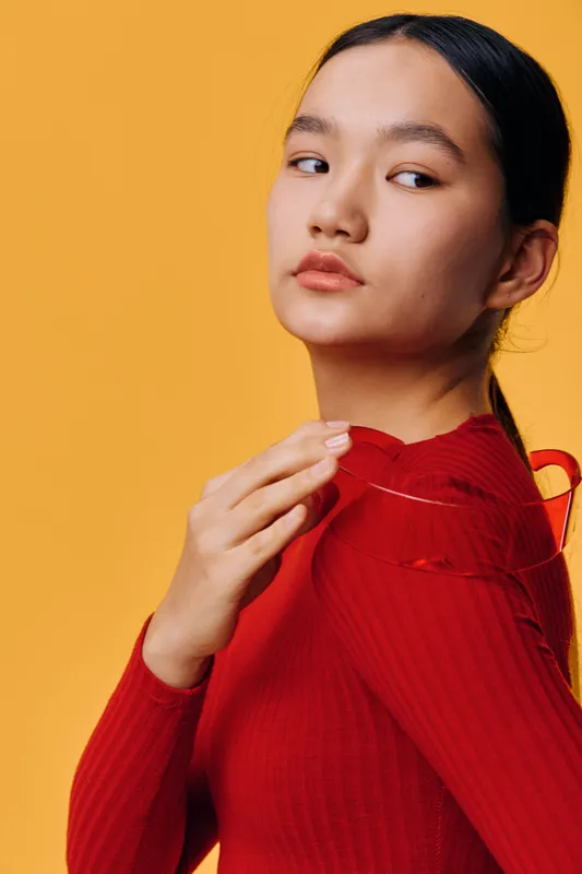 Portrait of young woman with dark hair and natural makeup wearing red sweater on orange background. Closeup of thoughtful female model looking sideways with hand on shoulder for fashion or beauty.