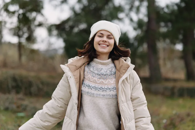 Woman in a warm coat and knit sweater standing outdoors, smiling in a chilly park with trees, capturing casual winter style and carefree outdoor mood.
