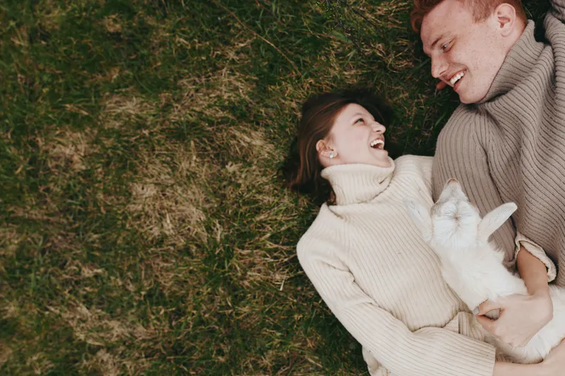 Couple lying on grass in cozy sweaters share a joyful moment with a fluffy rabbit, capturing intimate lifestyle romance outdoors and candid happiness under soft daylight