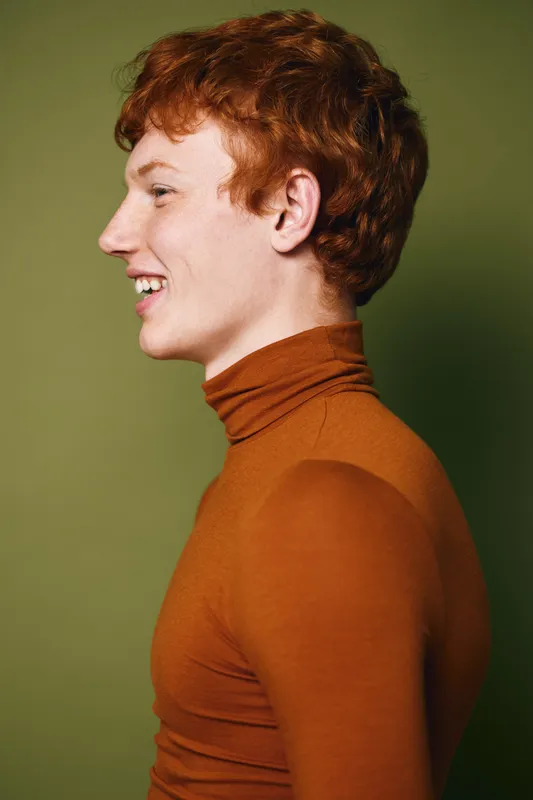Young man with red hair smiling and looking sideways on green background. Portrait of cheerful person wearing brown turtleneck standing against plain wall.