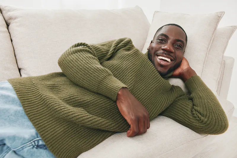 Smiling Black man relaxing on a beige couch, wearing a green sweater, showcasing happiness and comfort in a cozy indoor setting