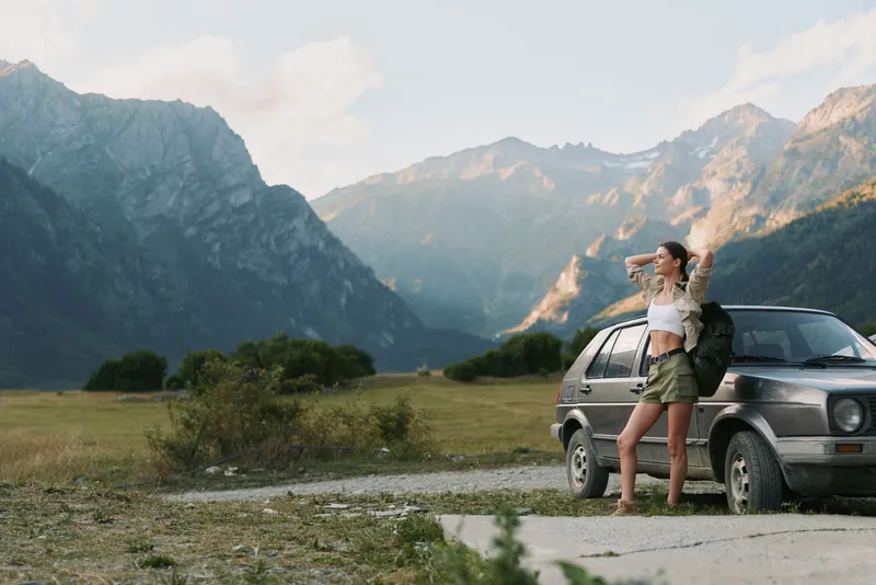 Young woman with backpack stands beside a car in a mountain valley, enjoying a bright sunlit moment and the expansive natural space around her.