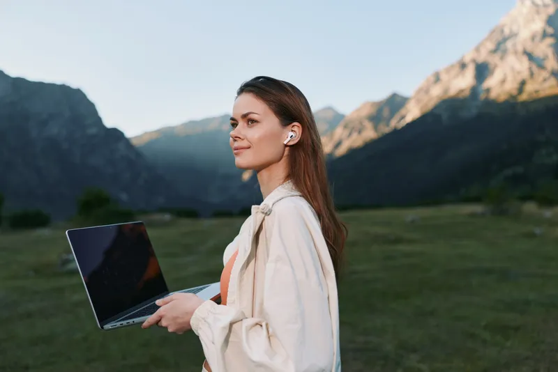 A professional woman stands outdoors in a scenic mountain landscape, holding a laptop and looking ahead with confidence, embodying focus, innovation, and modern work in nature.