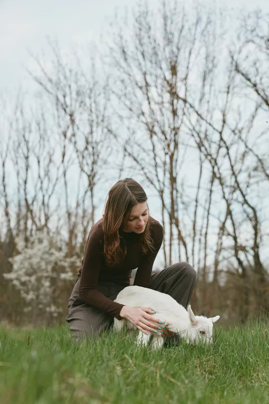 Young woman with long brown hair crouches in a grassy field gently petting a small white goat during a calm spring day with bare trees in the background.
