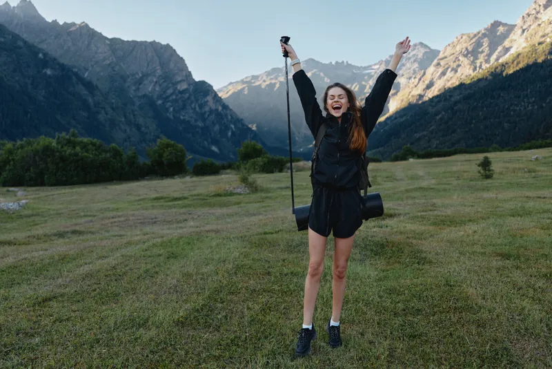 A cheerful hiker stands in a scenic valley, arms raised in triumph, smiling brightly as a camera bag rests at her side, capturing a moment of adventure and joy.