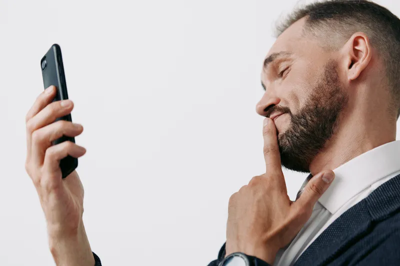 A professional business man stands against a colored isolated background, conveying thoughtful focus while reviewing messages on a smartphone.