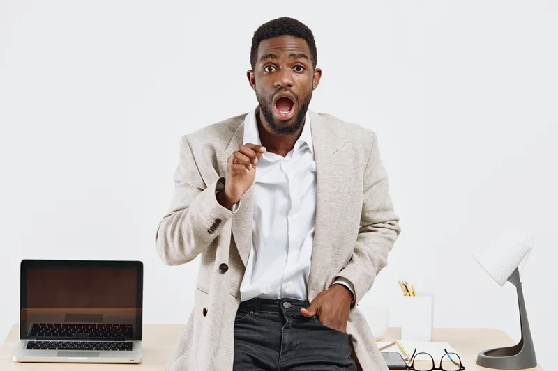 Concerned man in business casual attire stands at desk with laptop and office supplies, expressing surprise or shock with open mouth and wide eyes, isolated on plain white background. People lifestyle