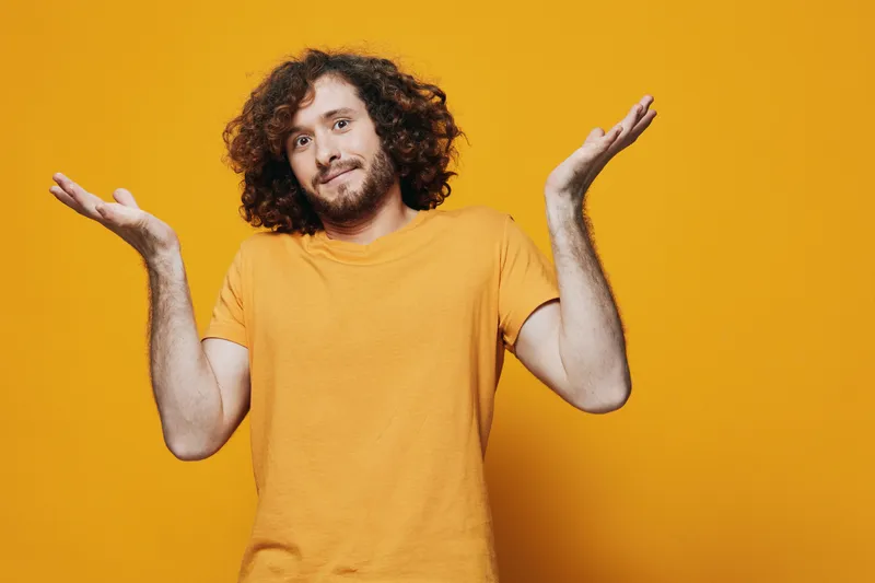 Cheerful young man with curly hair wearing an orange t shirt poses against a bright yellow background, expressing confusion and curiosity