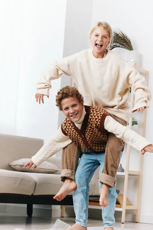 A playful couple indoors shares a piggyback moment with the woman riding on the man's back, both smiling broadly in a bright, modern living room.