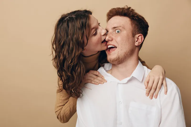 Young woman in brown sweater kisses surprised man in white shirt on cheek. Couple shows affection and playful emotions on beige background. Concept of love, relationship, and intimacy.