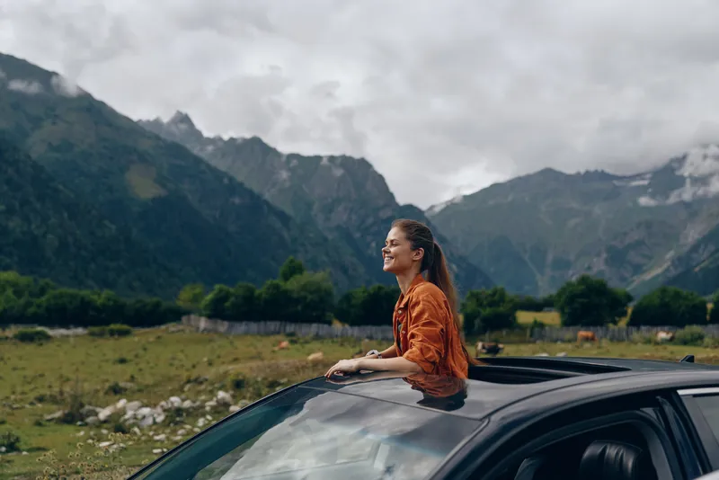 Woman leans from car window, smiling, enjoying an outdoor adventure in a mountainous landscape with open fields and dramatic clouds overhead under blue skies