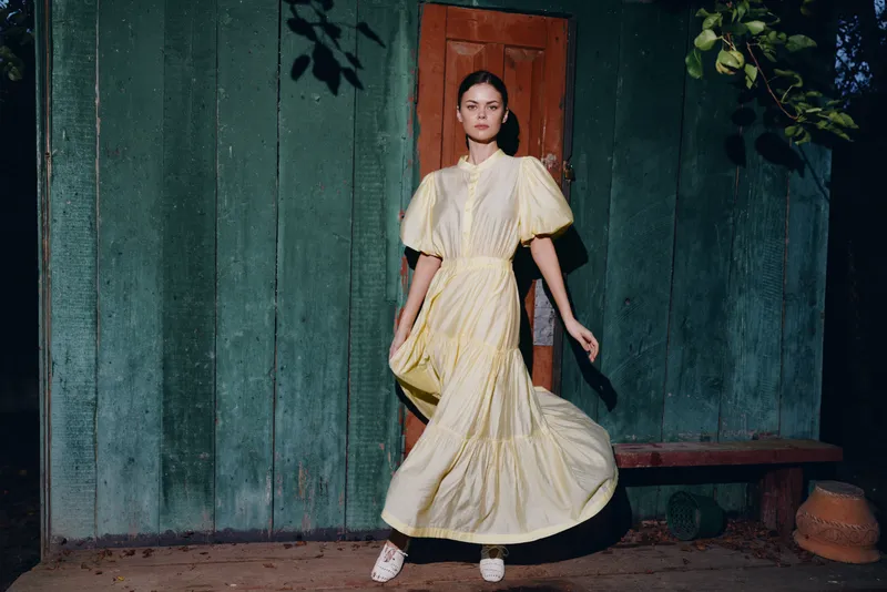 Woman in a flowing yellow dress poses outdoors beside a green wooden wall, creating a moody fashion moment under dusk lighting