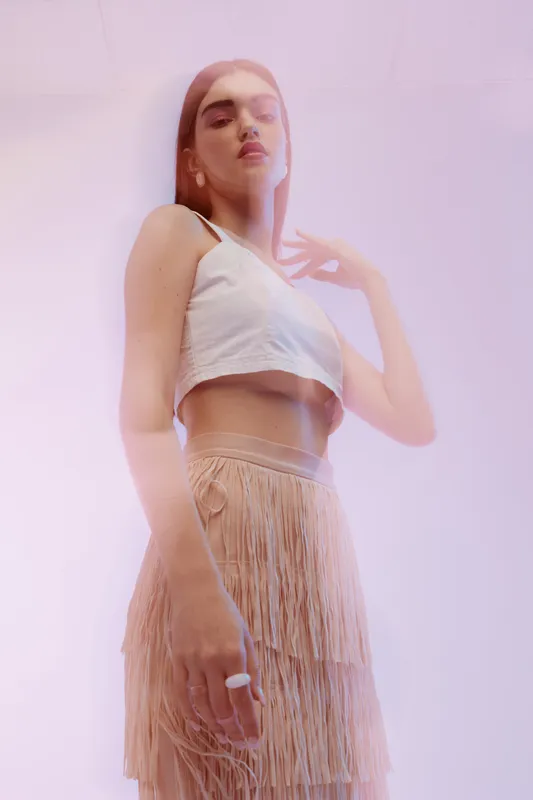 Young woman beauty fashion model posing in a white crop top and fringed skirt. Soft pink lighting creates a dreamy mood. Concept for style, elegance, and modern trends.