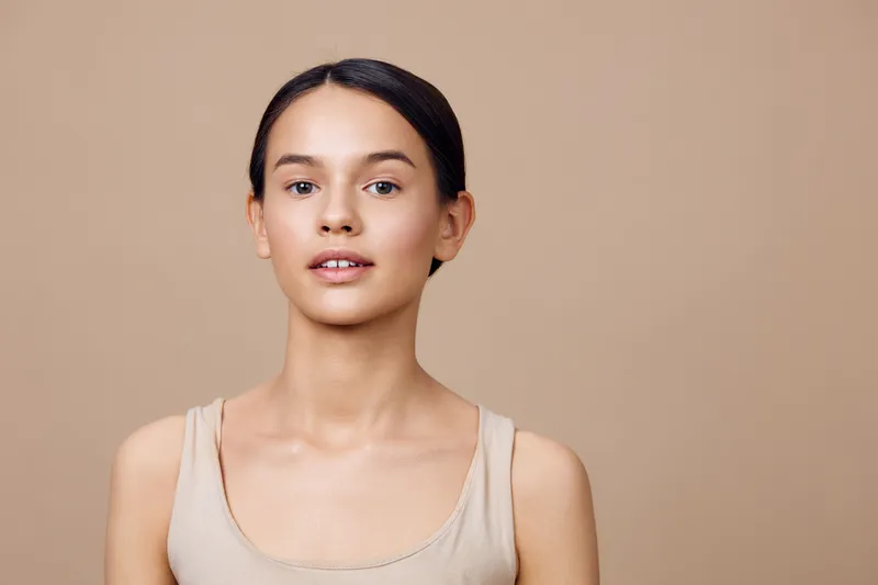 Young woman with natural beauty expressing confidence against a neutral background, showcasing minimal makeup and a casual outfit, highlighting healthy skin and simplicity