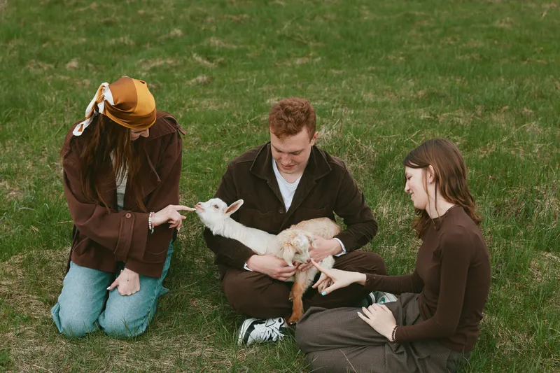 Three young adults enjoy a relaxed moment outdoors while holding and petting a small white baby goat on a grassy field during a casual lifestyle gathering.