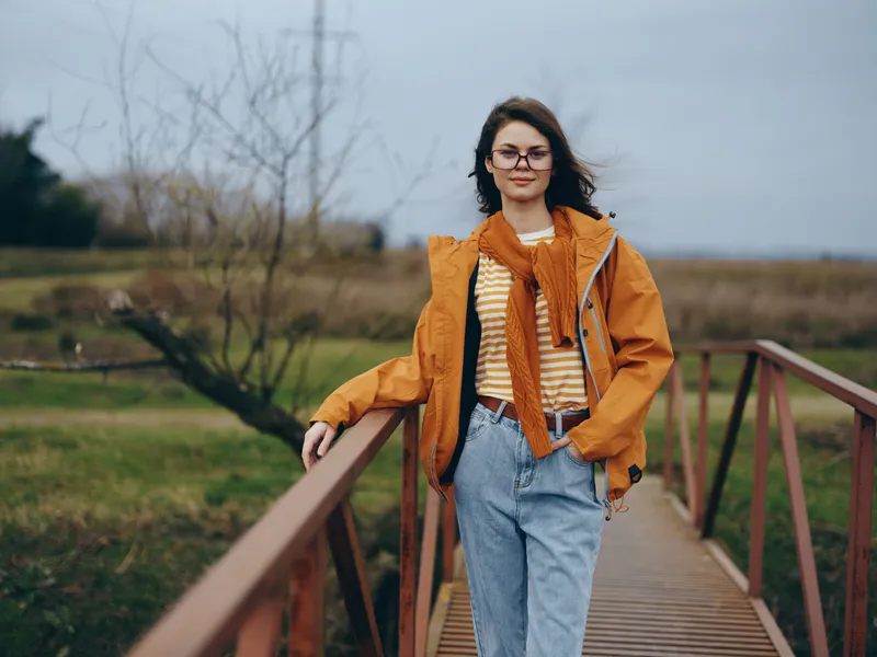 Woman lifestyle outdoors on a wooden bridge, wearing orange jacket and jeans, walking in a rural park, casual spring mood, cloudy sky, natural light, relaxed pose, background trees today