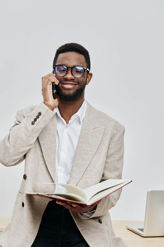 Cheerful young man with glasses in smart casual outfit talking on mobile phone while holding an open book, sitting at desk with laptop against plain white background, cheerful expression. People lifes