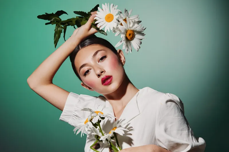 Beauty creative portrait of a young woman with smooth skin and bold red lips holding daisies. Fashion concept with studio lighting on a green background.