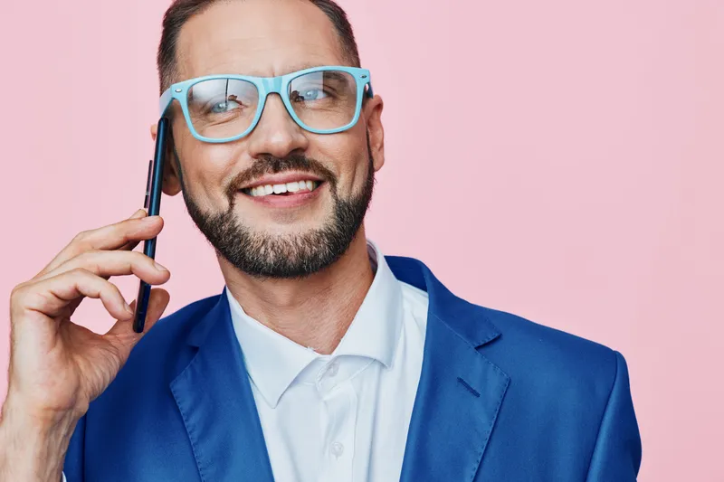 A professional businessman on a solid pink backdrop, smiling while talking on a phone, embodying confidence, optimism, and forward thinking in a modern work setting.