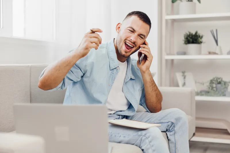 Excited man communicating on a phone, wearing casual attire with a joyful expression, surrounded by a modern, bright workspace, showcasing productivity and creativity.