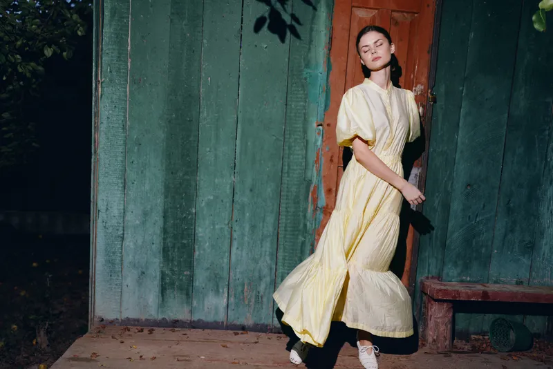 Portrait of a woman model in a flowing yellow dress stands by a weathered doorway in an outdoor night setting, striking a poised pose and glancing back, exuding elegant fashion and moody cinematic