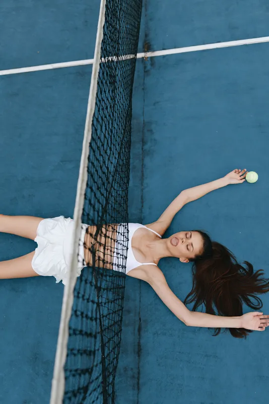 Young female tennis player resting on a blue court, wearing a white athletic outfit, embodying strength and determination with an expression of focus