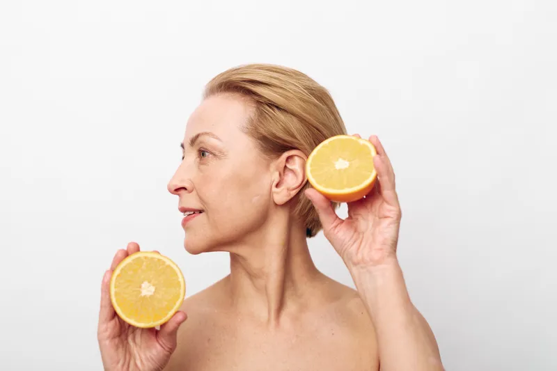 Middle aged woman with short blonde hair holds two halves of fresh orange near face on white background. Concept of healthy lifestyle, natural skincare, and vitamin c benefits.