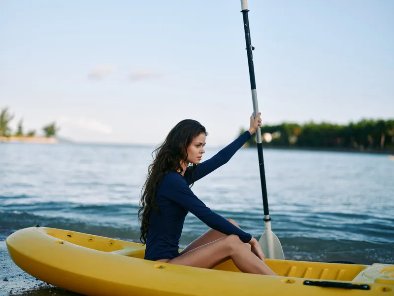 A young woman sits in a yellow kayak on calm water, gripping a paddle and gazing toward the horizon as soft light highlights her contemplative pose.