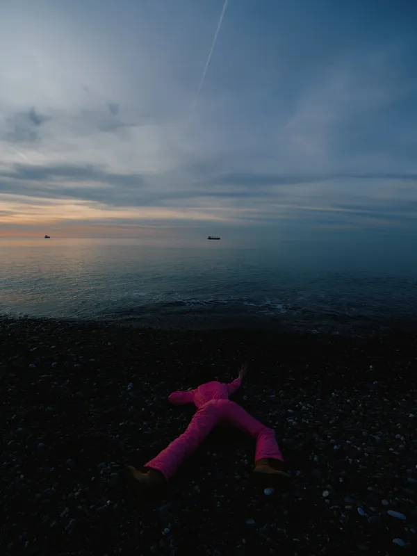 Beach scene at sunset with a person lying on the shoreline in a pink outfit, facing the calm sea and distant boats, creating a tranquil contemplative mood for viewers and photographers alike