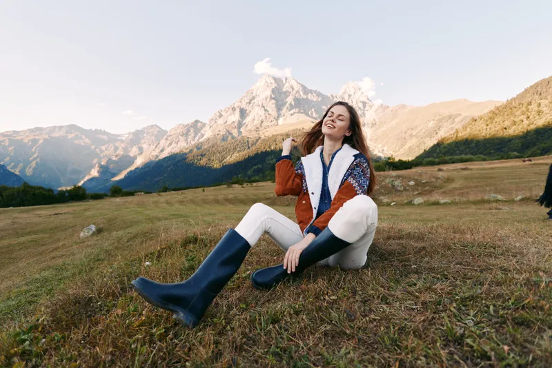 Woman sitting on mountain meadow smiling in colorful jacket and rubber boots, relaxed travel portrait in nature with distant peaks, outdoor hiking mood and warm sunlight.