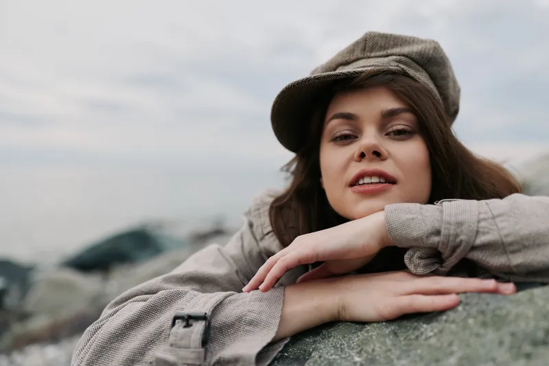 Young woman in casual clothes and cap rests on rock outdoors, looking relaxed and confident. Overcast sky and natural background create calm, contemplative mood and outdoor lifestyle concept.