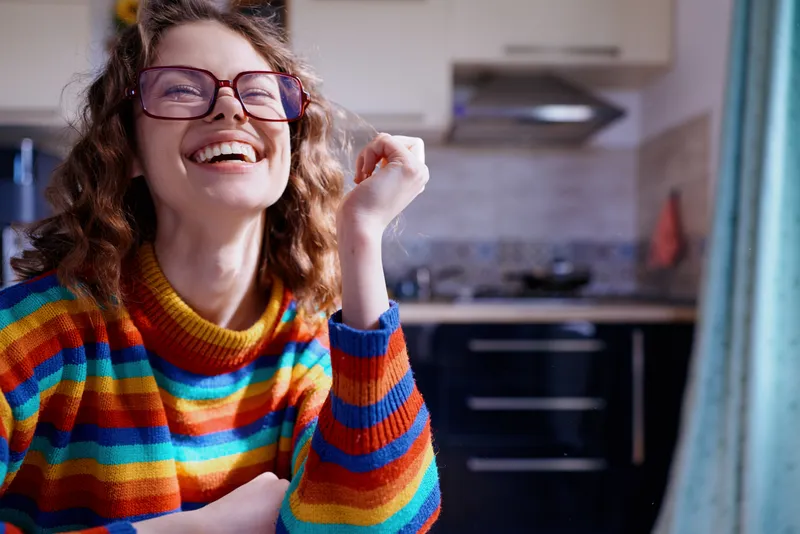 Joyful woman, smile, laughter, at home in a colorful striped sweater, standing in a bright kitchen, casual lifestyle moment with cozy interior and warm light
