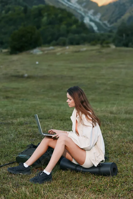 A focused woman sits on a grassy meadow, typing on a laptop in an outdoor setting. She wears a light jacket and sneakers, projecting a calm, productive and modern work vibe.