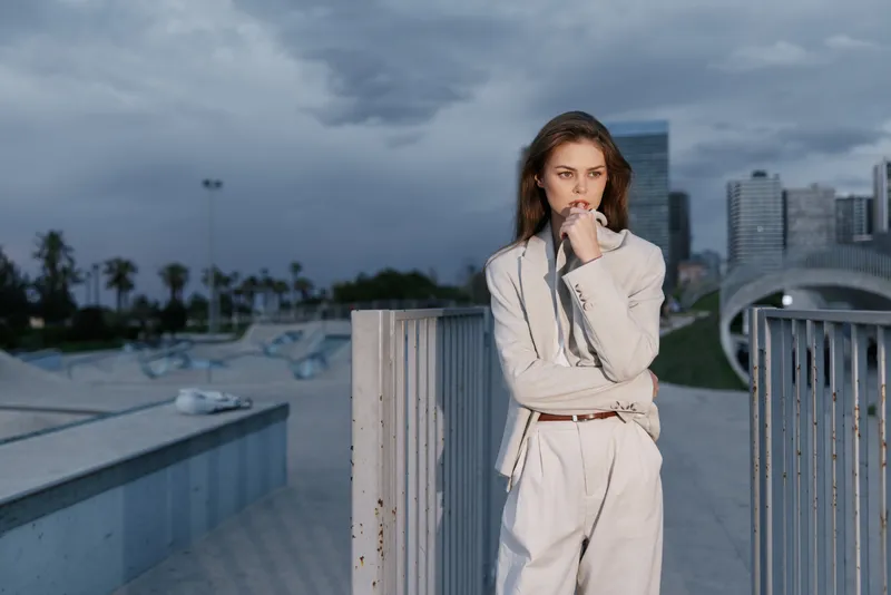 Woman in white suit thinking outdoors with cityscape and cloudy sky, businesswoman looking focused and determined near urban skate park