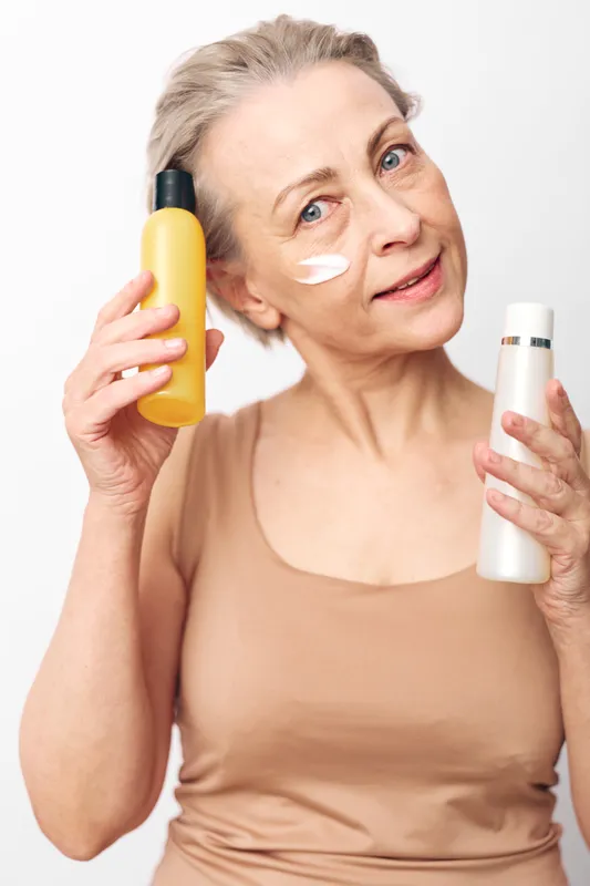 Mature woman with light skin applies face cream with a smile, holding two cosmetic bottles. Concept of skincare routine, anti aging, wellness, and natural beauty with soft lighting.